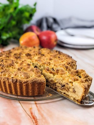 Apple crumble tart served on plate with one piece on a spatula and apples in the background.