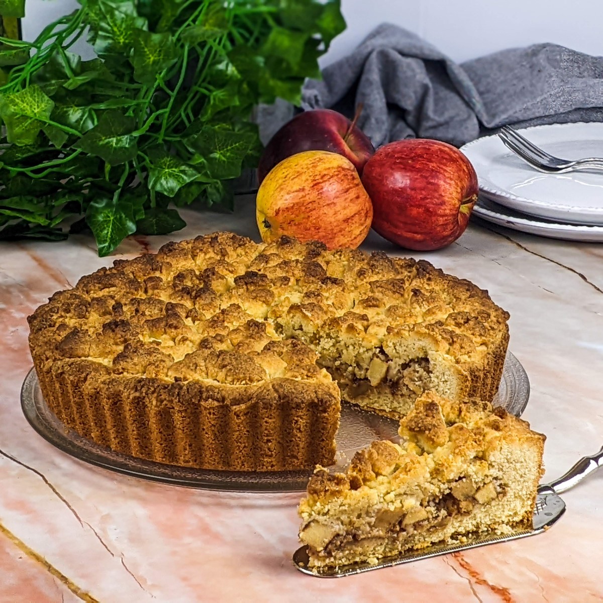Apple crumble tart served on plate with one piece on a spatula and apples in the background.