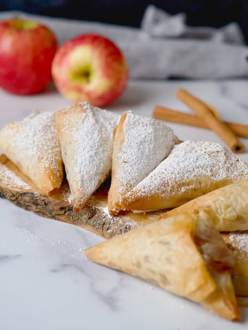 Apple turnovers on a wooden board with powdered sugar on top and some apples in the back.