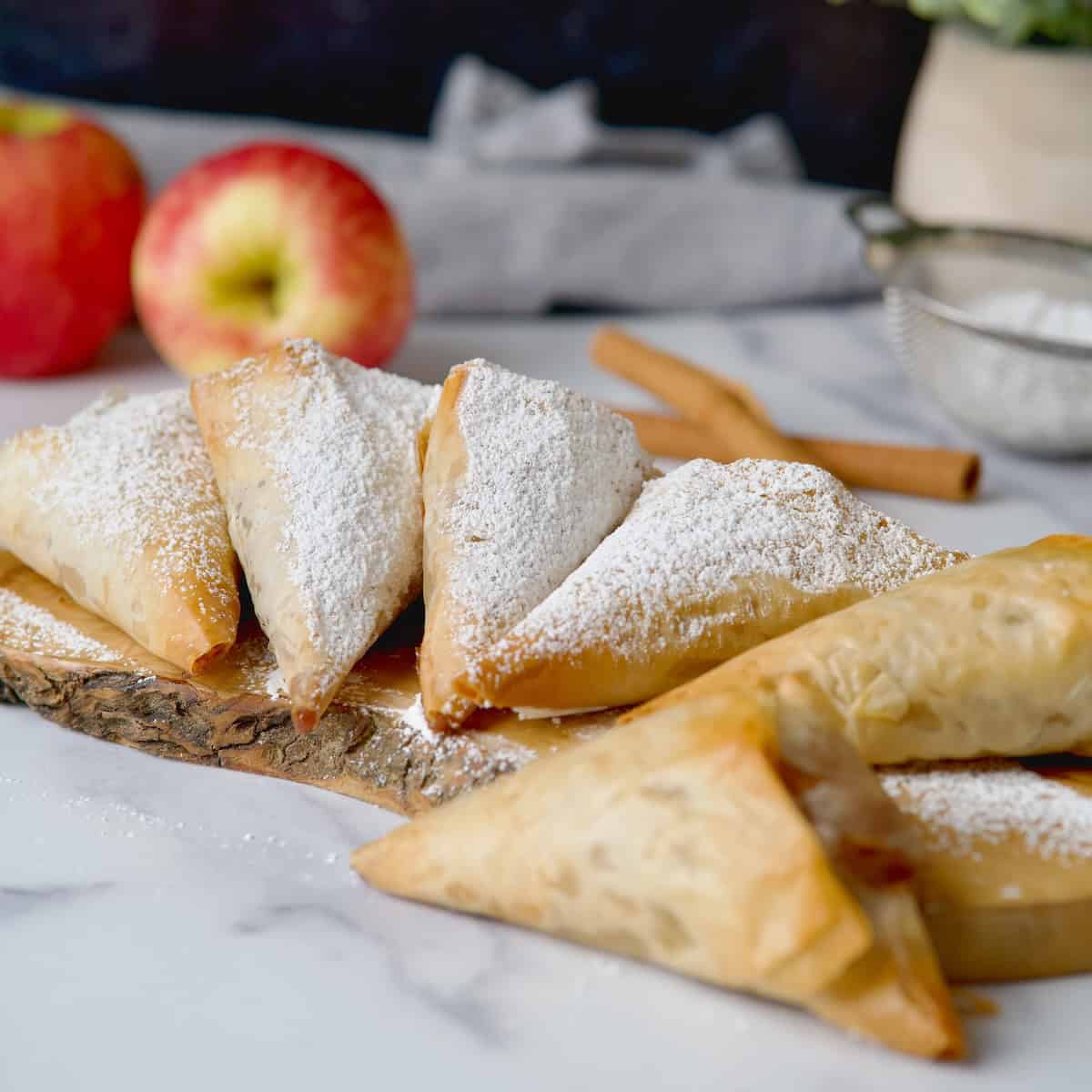 Apple turnovers on a wooden board with powdered sugar on top and some apples in the back.