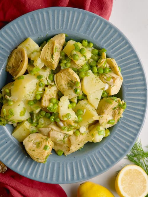 Artichoke and potato salad served in a blue bowl next to a lemon and a jar with olive oil.
