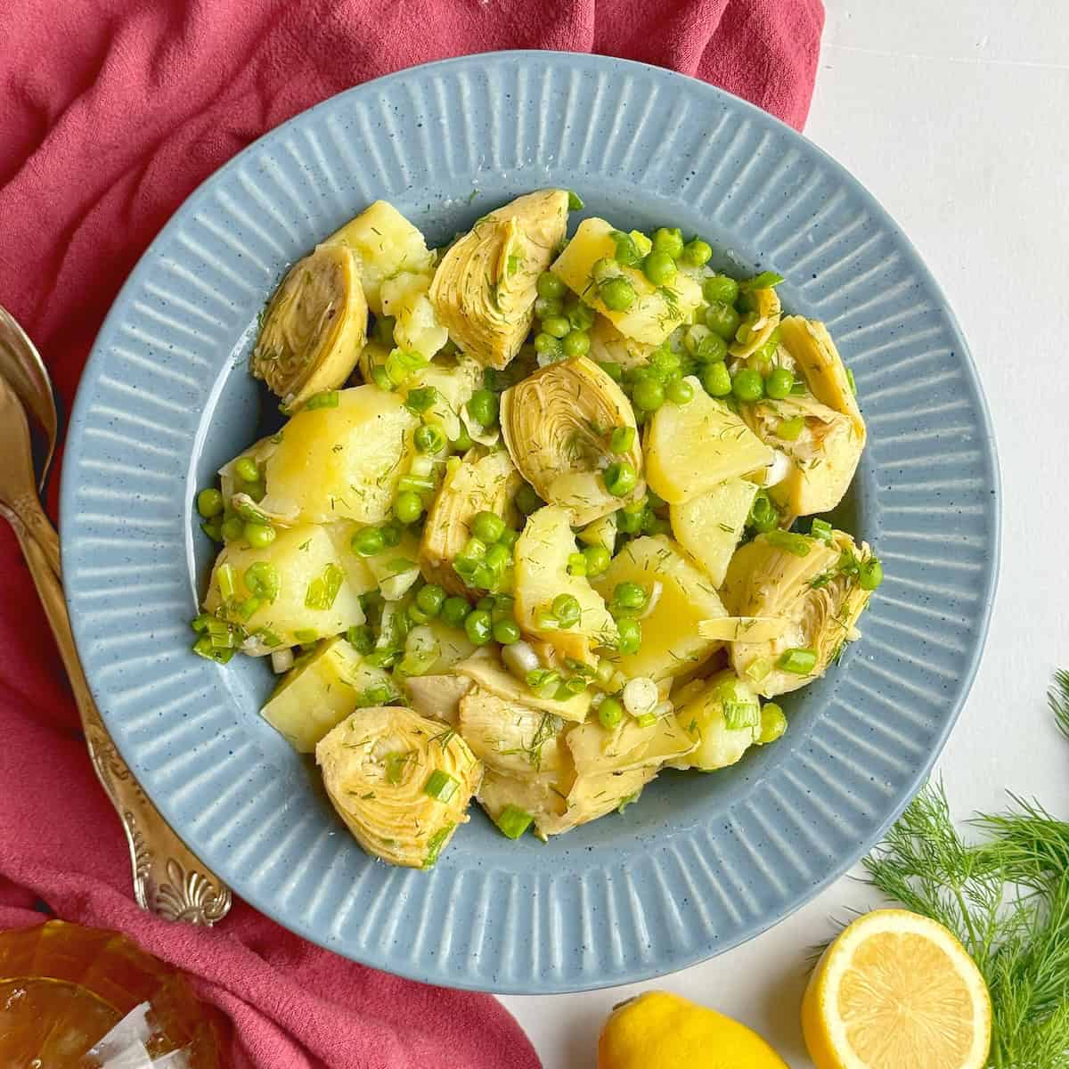 Artichoke and potato salad served in a blue bowl next to a lemon and a jar with olive oil.