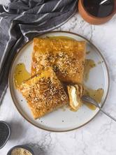 Two blocks of baked feta wrapped in filo served on plate with a bowl of honey and sesame seeds next to it.