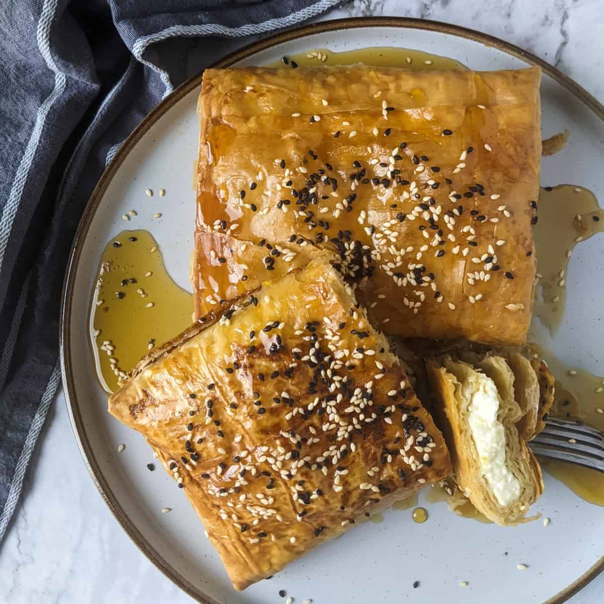 Two blocks of baked feta wrapped in filo served on plate with a bowl of honey and sesame seeds next to it.
