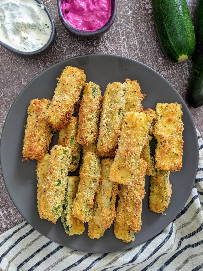 Zucchini fries on grey plate next to yoghurt and beetroot dip and two zucchinis