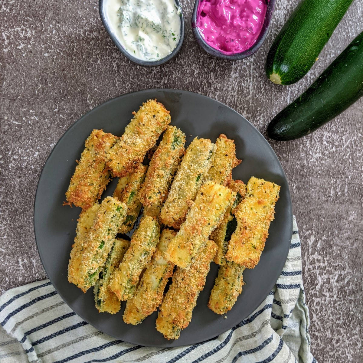 Zucchini fries on a gray plate next to bowl with yogurt dip and beetroot dip.