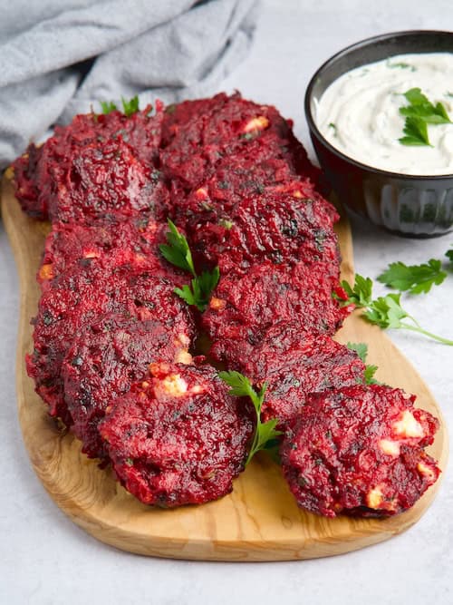 Beet fritters layed on a wooden board next to bowl with white sauce.