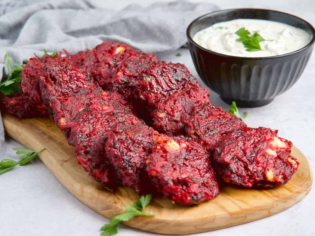 Beet fritters on a wooden board next to bowl with sauce.