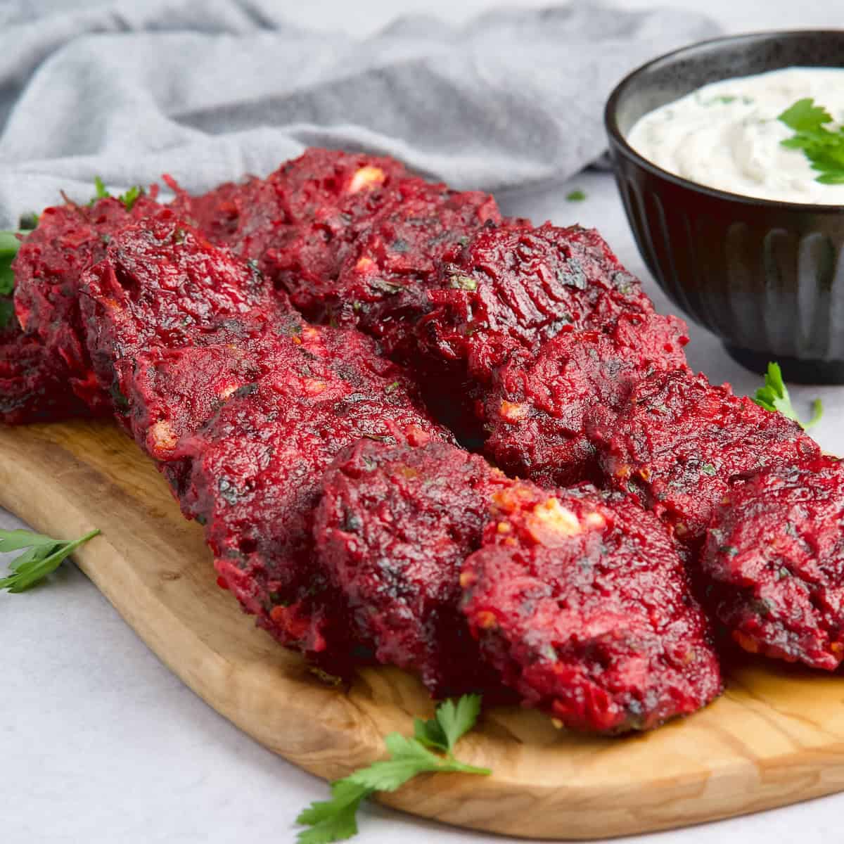 Beet fritters layed on a wooden board next to bowl with white sauce.