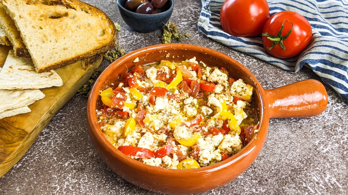 Baked feta cheese with tomatoes in an ovenproof dish next to bread, olives and tomatoes.