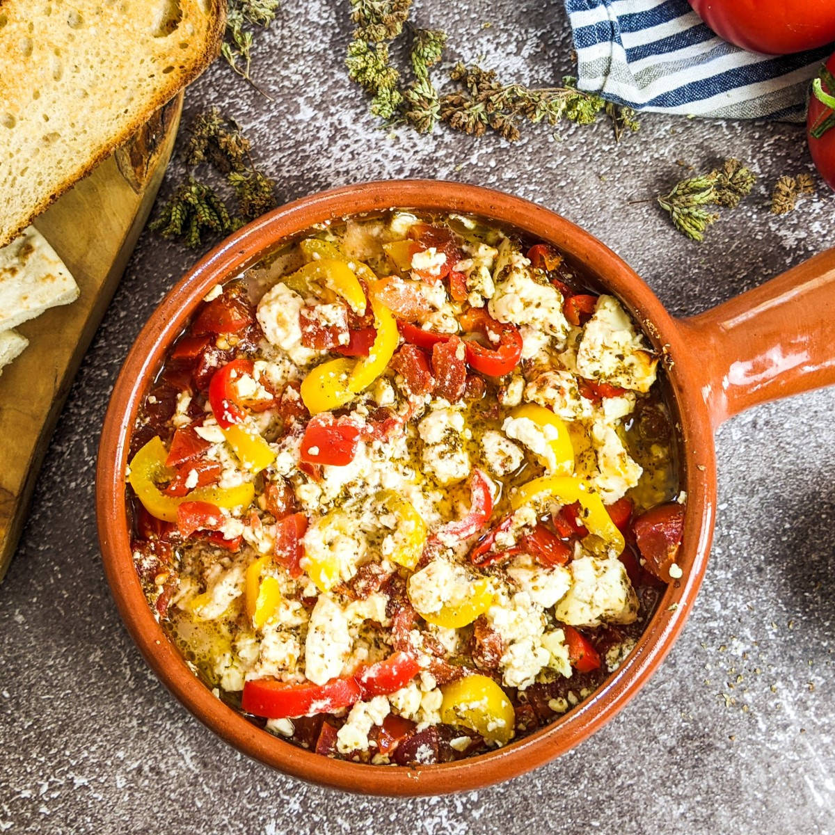 Baked feta cheese with tomatoes in an ovenproof dish next to bread, olives and tomatoes.
