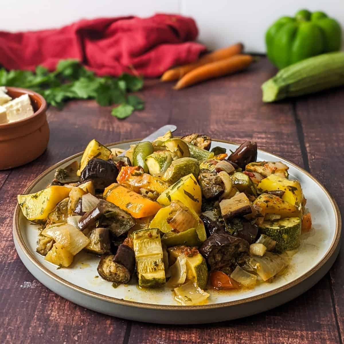 Briami casserole served on a plate next to raw vegetables and a bunch of parsley.