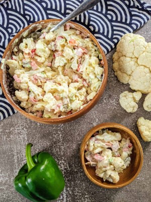 Cauliflower salad served in wooden bowl next to cauliflower florets and pepper.