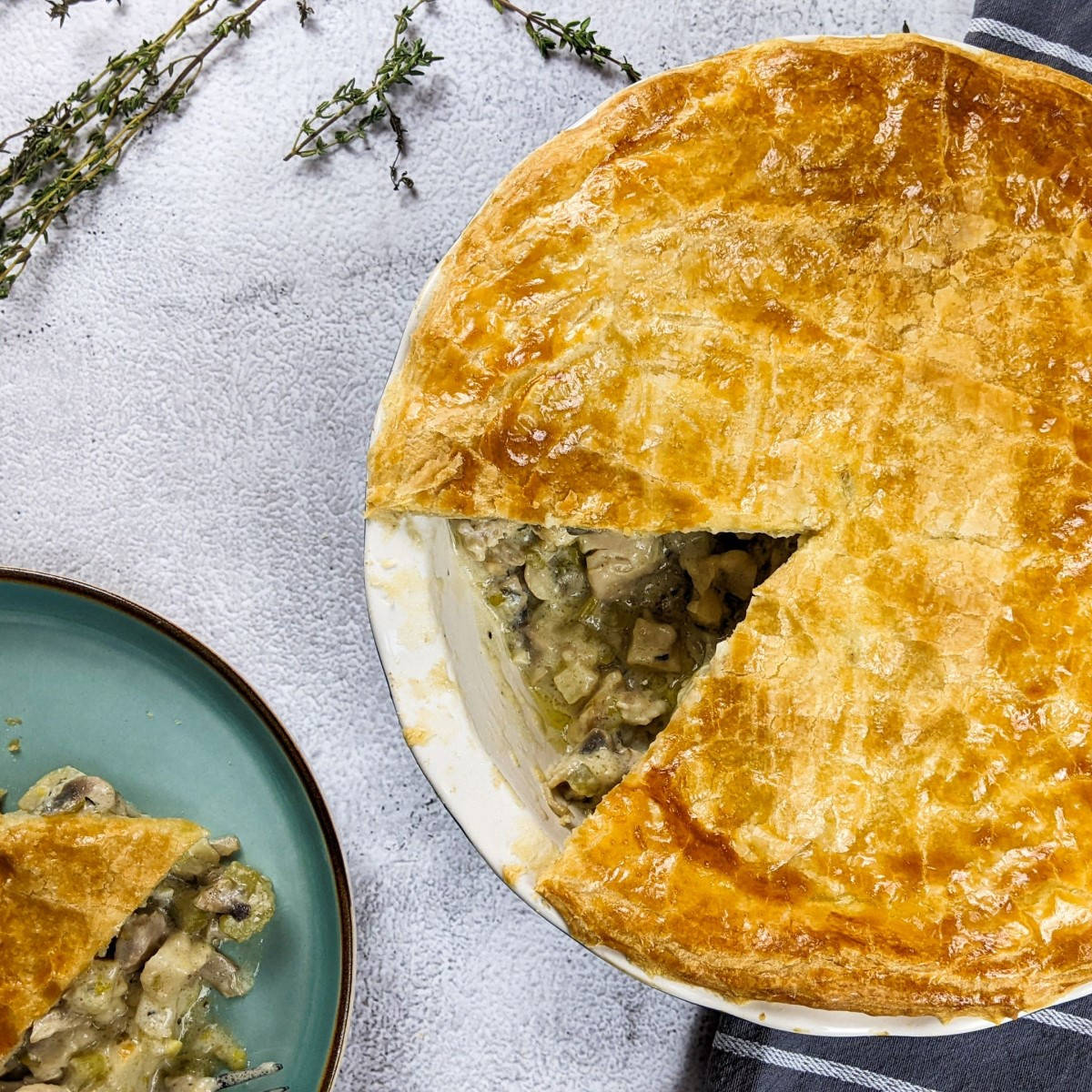 Celery, mushroom and chicken pie slice with the whole pie dish next to it and some thyme and mushrooms.