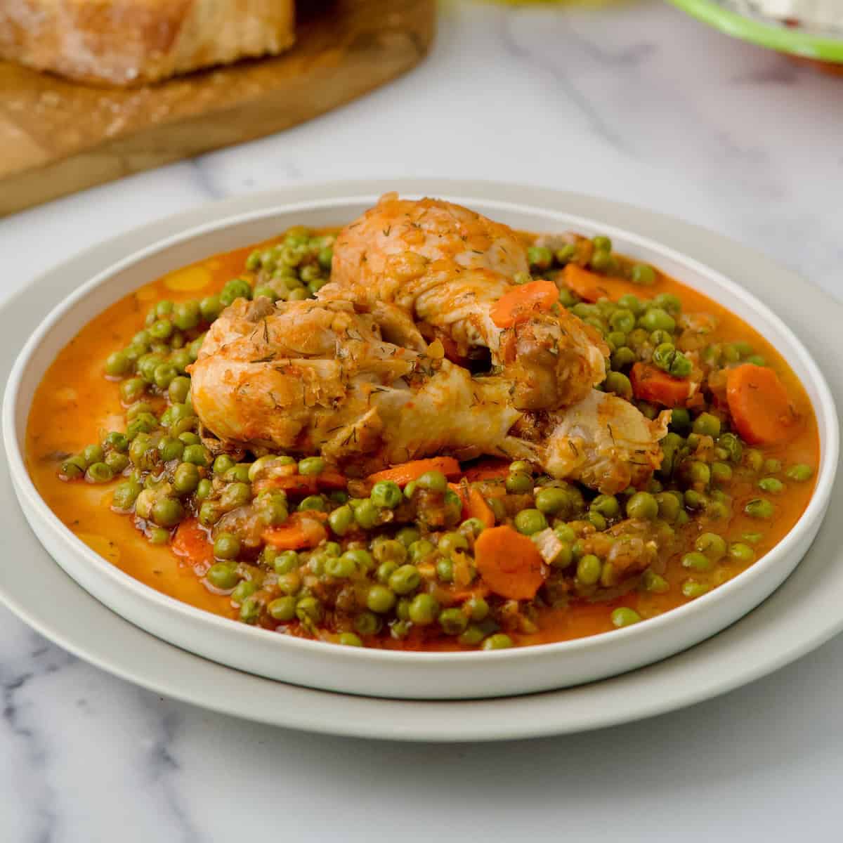 Chicken with green peas served on white plate with slices of bread in the background.