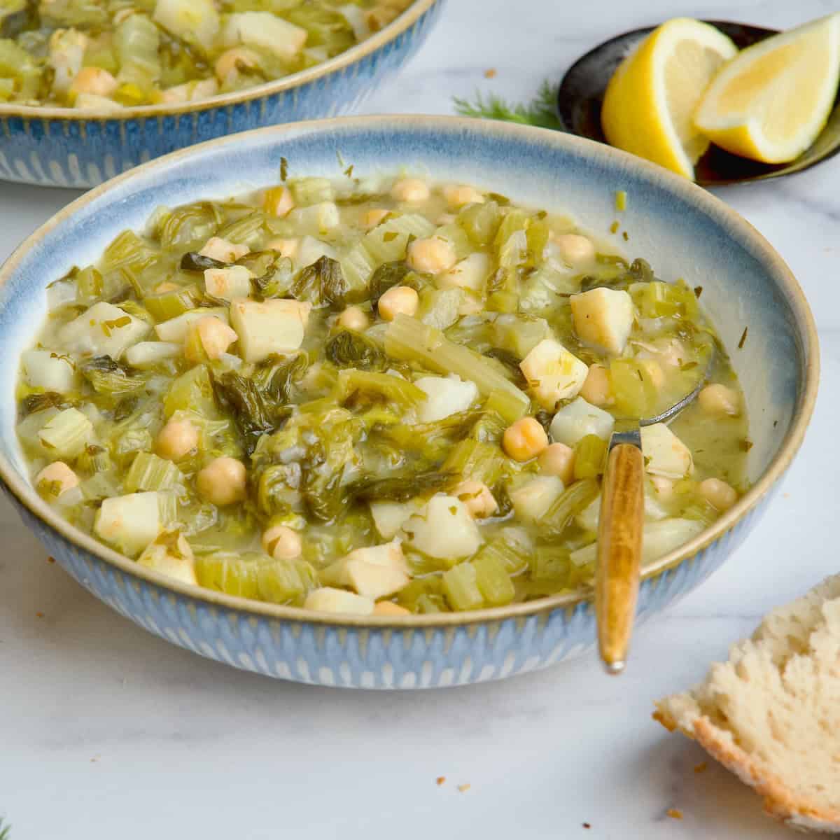 Chickpea and celery stew served in white bowl next to loaf of bread and lemon slices.