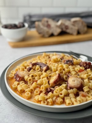 Octopus pasta served on white plate with bread slices and olives in the background.