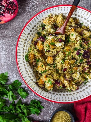 Mediterranean couscous salad with squash served in a bowl next to parsley, pomegranate and a bowl with raw couscous.