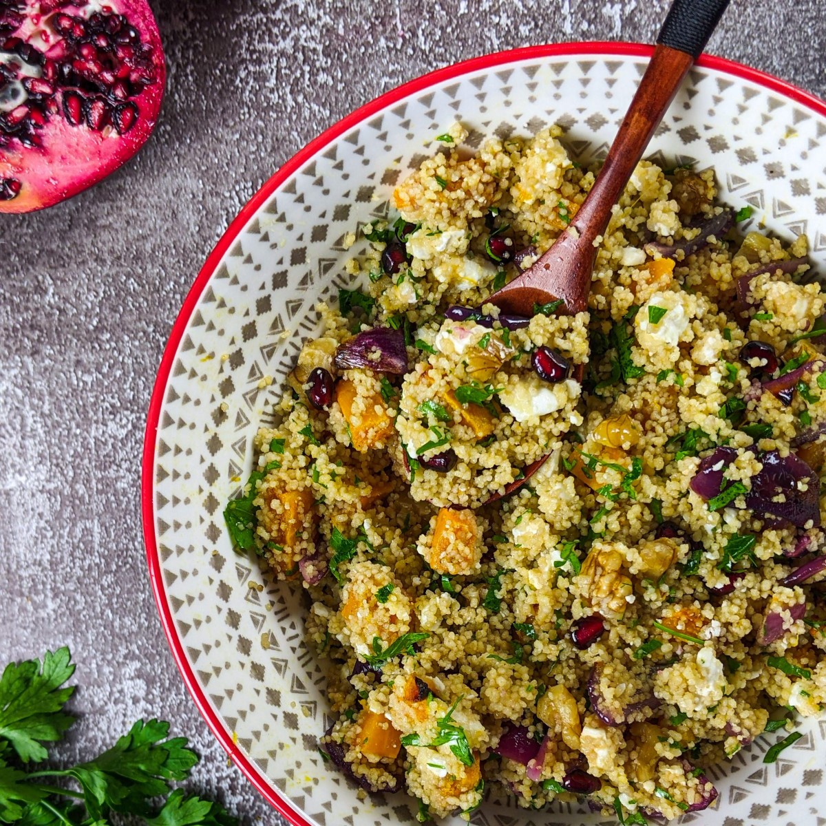 Mediterranean couscous salad with squash served in a bowl next to parsley, pomegranate and a bowl with raw couscous.