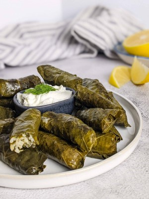 Stuffed grape leaves on a plate next to a bowl with yogurt and lemon slices.