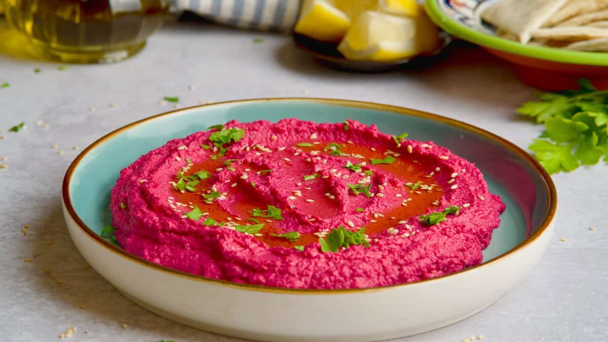Beet hummus on a blue plate with sesame seeds, oil and chopped parsley on top.