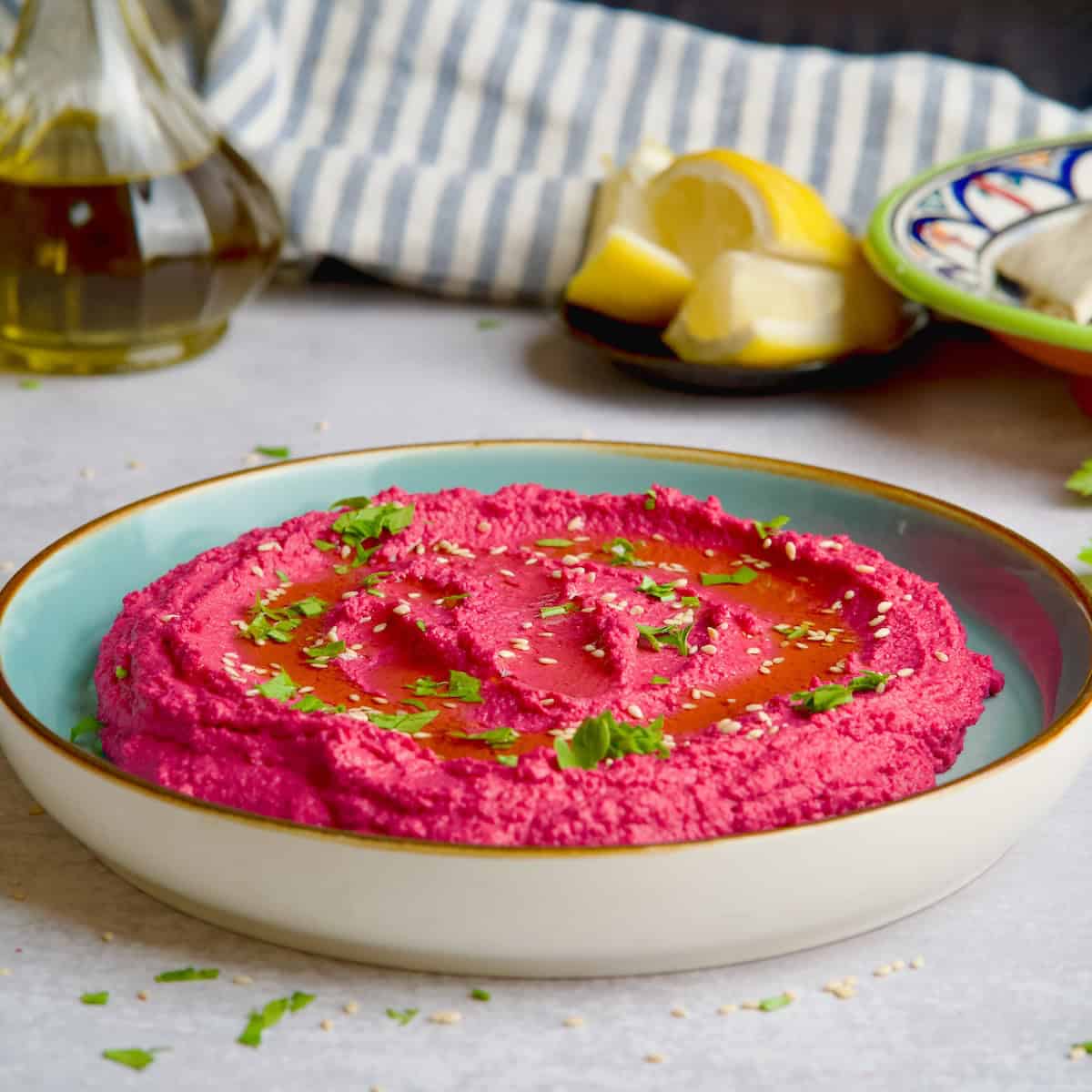 Beet hummus served on blue plate with sesame seeds, chopped parsley and oil on top.