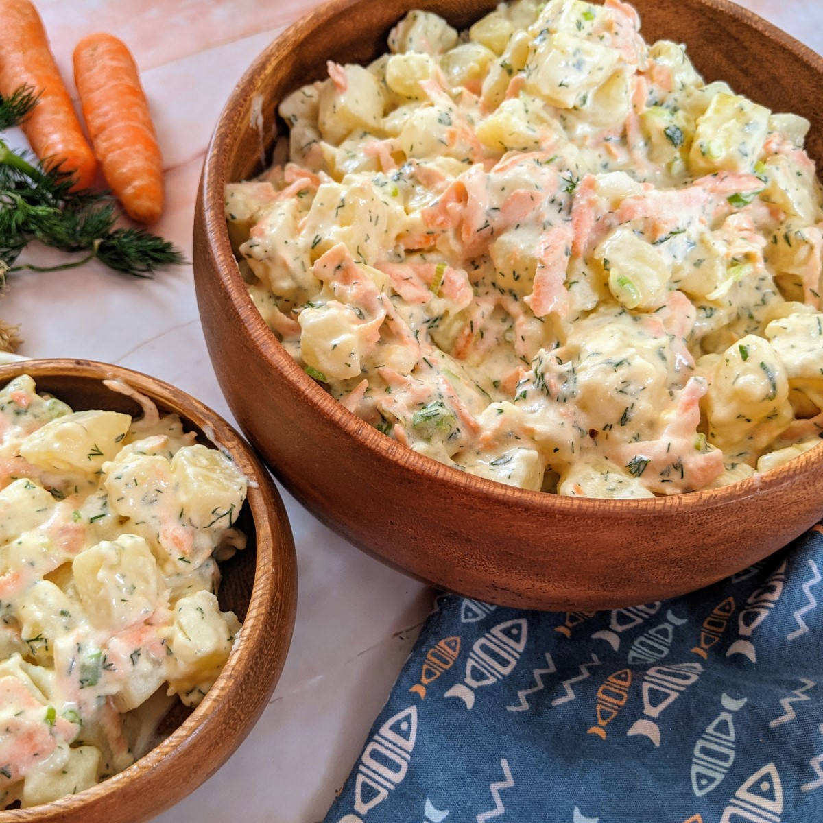 Creamy potato salad served in wooden bowl next to a carrot, green onions and potatoes.