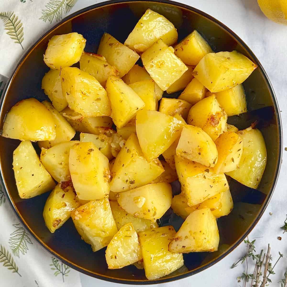 Greek lemon potatoes in a black plate next to half lemon and dried herbs.