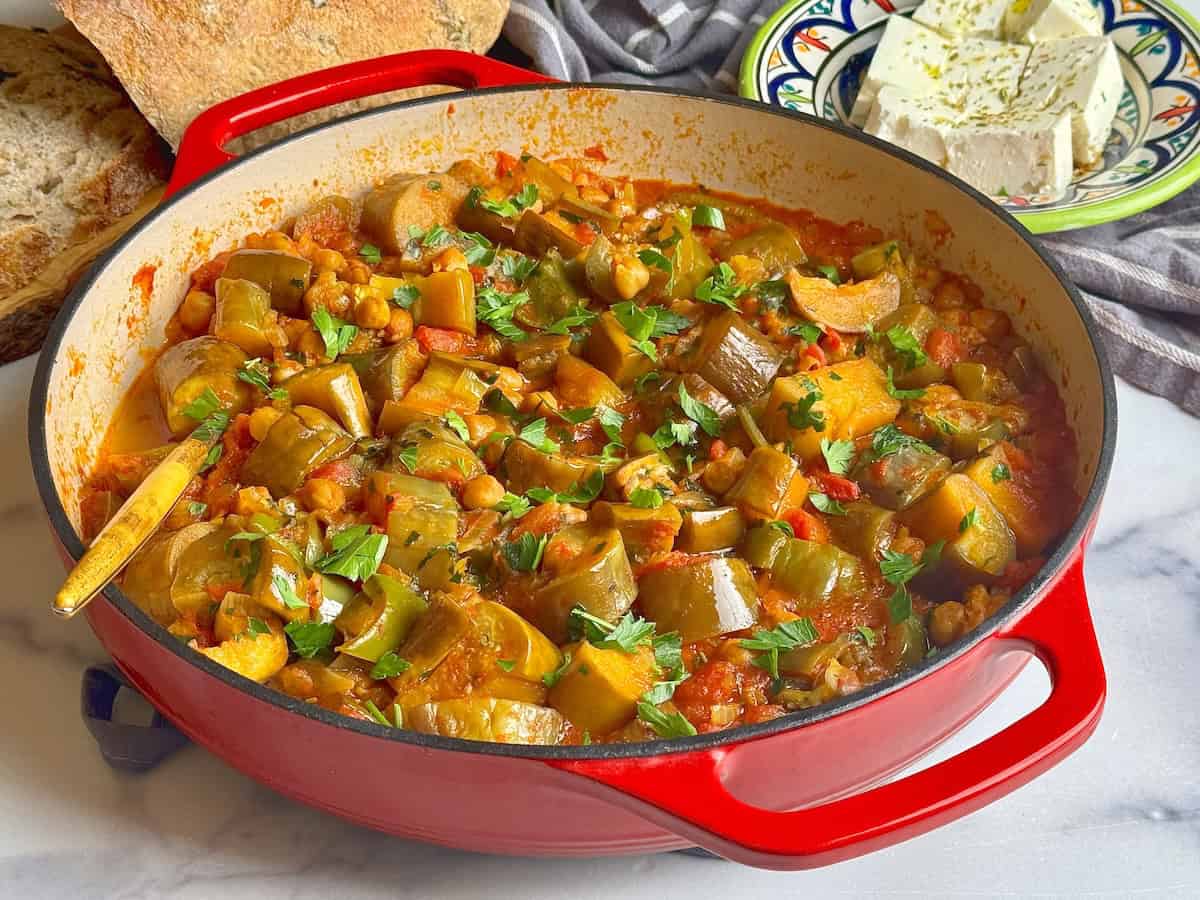 Chickpea and eggplant stew served in a red cast iron pan next to bread and feta cheese.