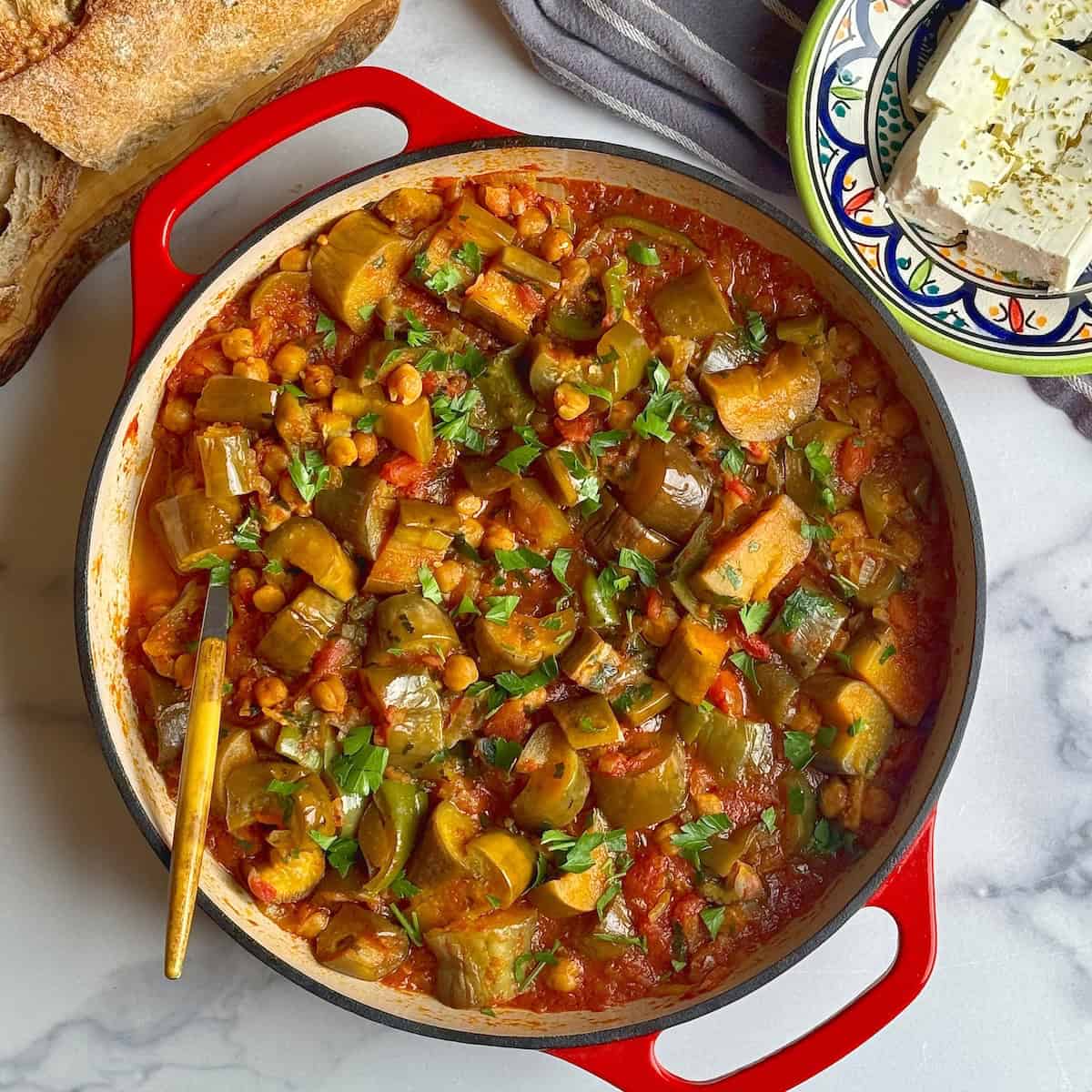 Eggplant and chickpea stew in a red cast iron pan next to plate with feta cheese and a loaf of bread.