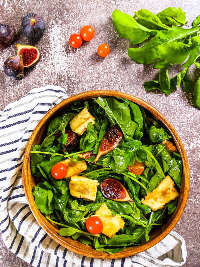 Fig and halloumi salad served in bamboo bowl next to tea towel, figs, cherry tomatoes.