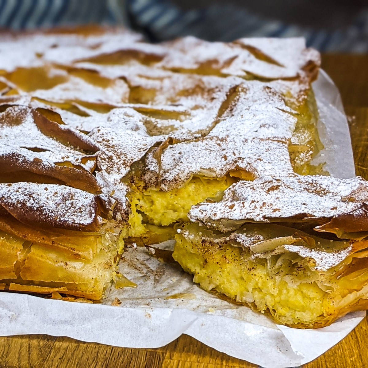 Greek custard pie with pumpkin dusted with powdered sugar and cinnamon on a wooden board and one slice cut.