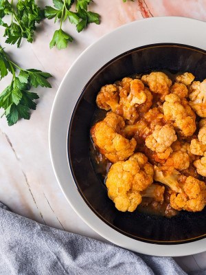 Tomato braised cauliflower served on a plate next to cauliflower pieces and fresh parsley. Tomato braised cauliflower served on a plate next to cauliflower pieces and fresh parsley.