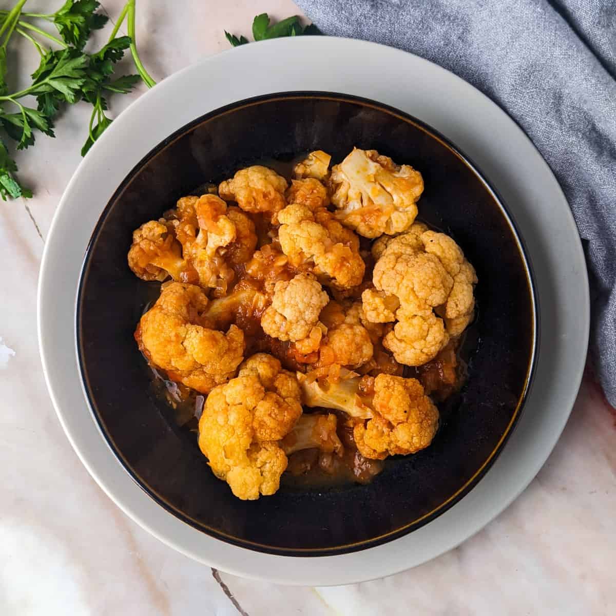Tomato braised cauliflower served on a plate next to cauliflower pieces and fresh parsley.