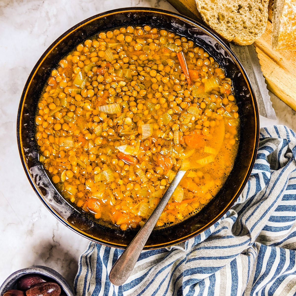 Mediterranean lentil soup served in black bowl next to olives and loaf of bread.