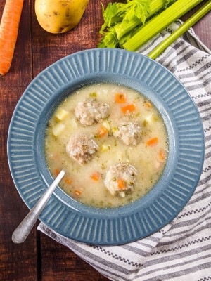 Greek meatball soup served in a blue bowl next to carrot, potato and celery.