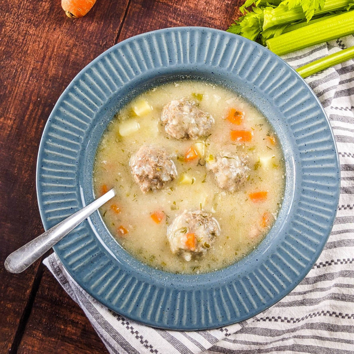 Greek meatball soup served in a blue bowl next to carrot, potato and celery.