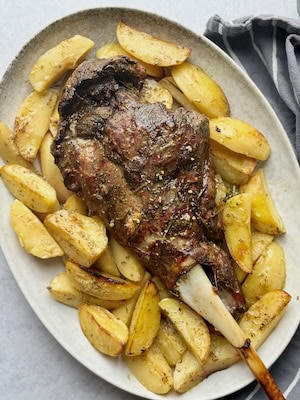 Roasted leg of lamb with potatoes around it in a white serving plate.