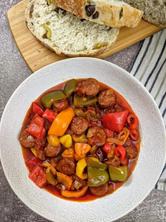 Greek sausages and peppers in tomato sauce served in beige bowl next to slices of bread