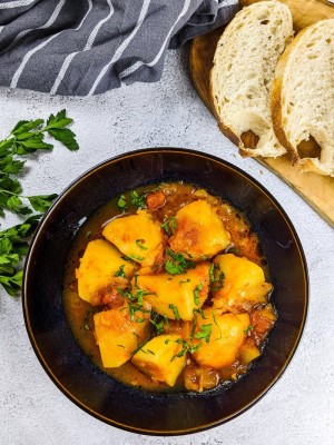 Greek tomato potatoes stew served in black bowl with chopped parsley on top and slices of bread next to it.