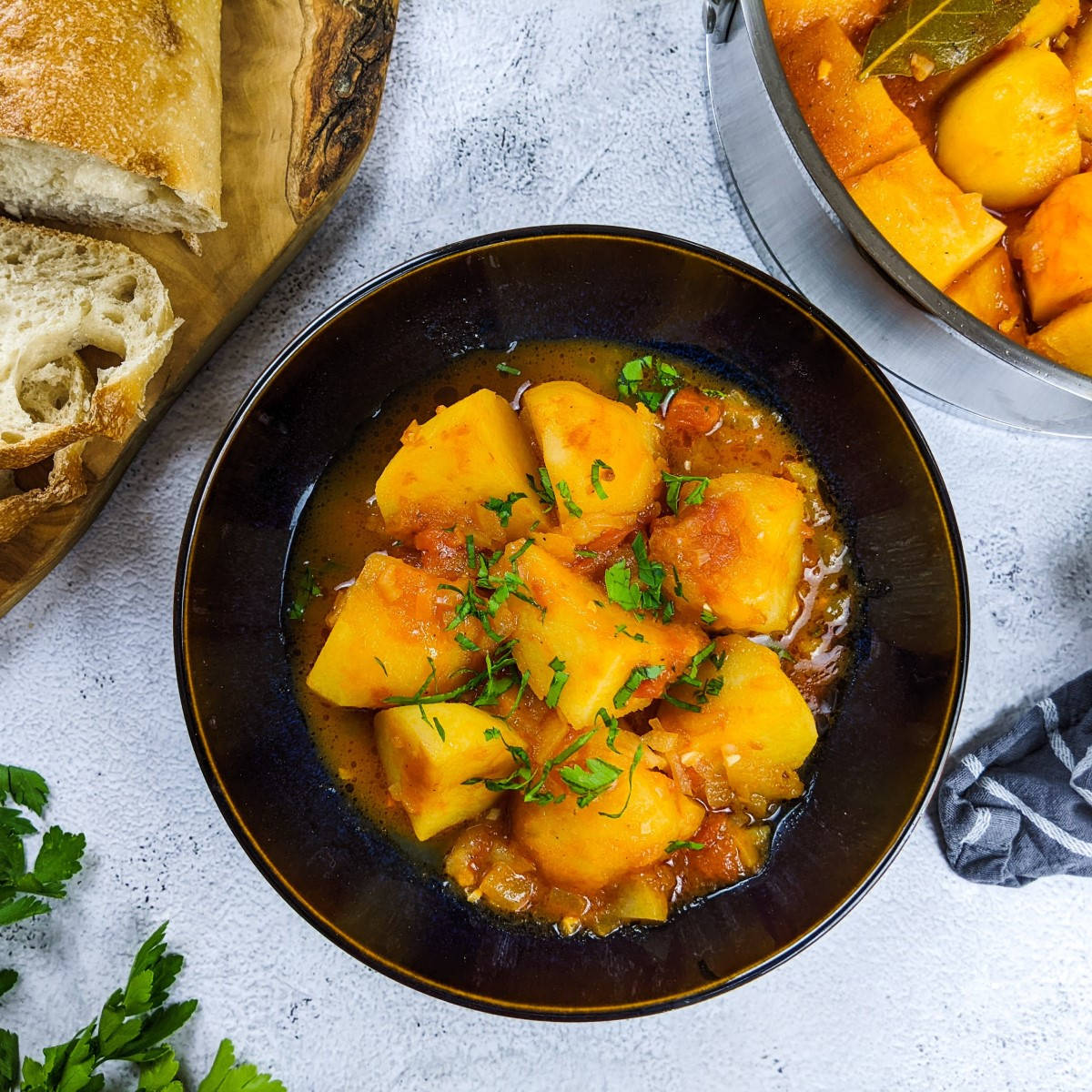 Greek tomato potatoes stew served in black bowl with chopped parsley on top and slices of bread next to it.