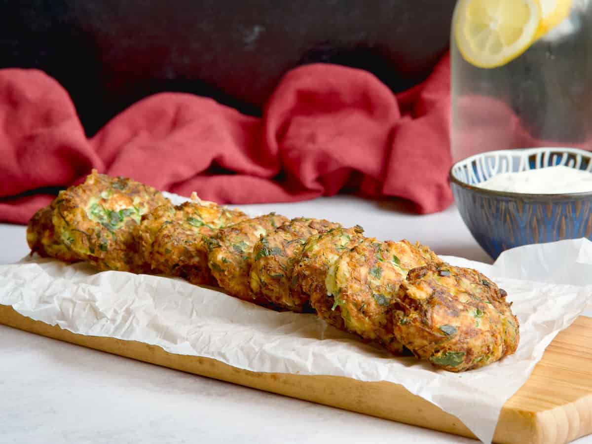 Zucchini fritters on a wooden board next to bowl with yogurt sauce.