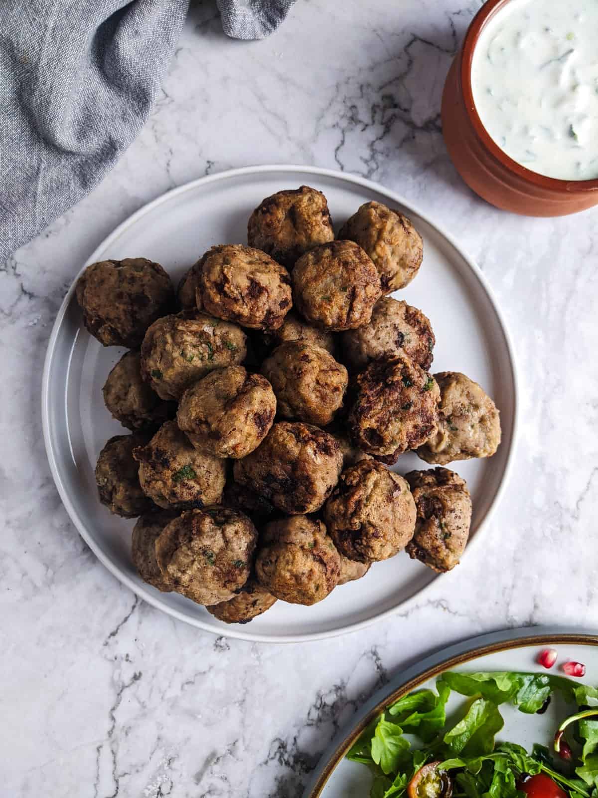 Meatballs served on white plate next to salad and bowl with tzatziki dip.