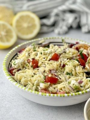 Orzo pasta and feta salad served in bowl next to lemon slices and orzo pasta.