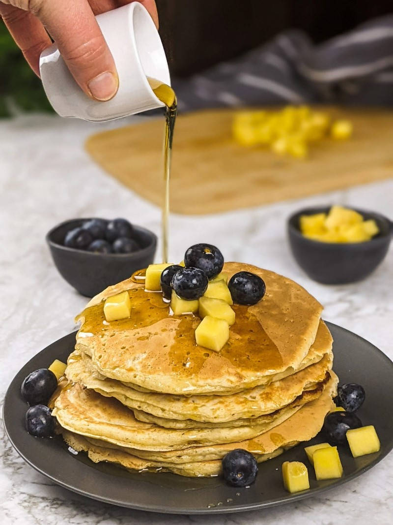 Pancakes το ένα πάνω στο άλλο με μύρτιλα και κομματάκια μάνγκο από πάνω και σιρόπι σφενδάμου να τρέχει.