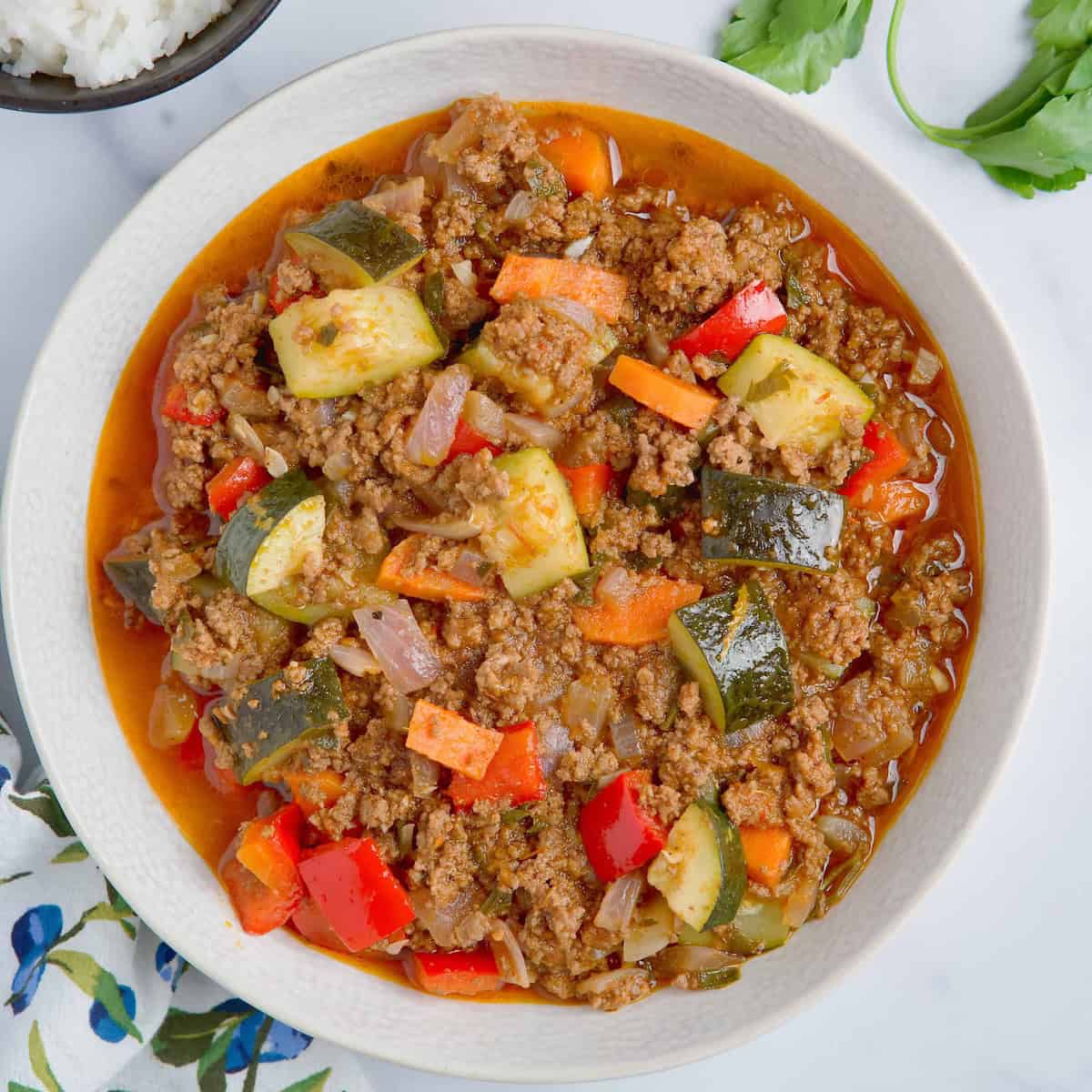 Ground beef and vegetables served in white bowl next to fresh parsley, rice and olive oil.