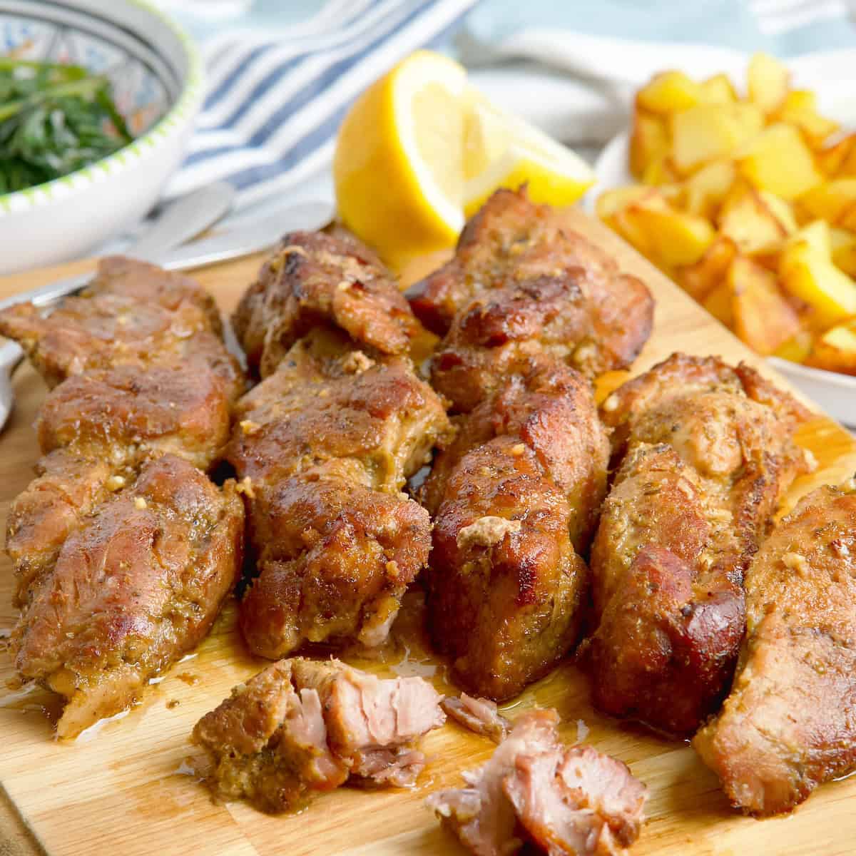 Pork shoulder ribs on a wooden board next to bowl with greens and potatoes.