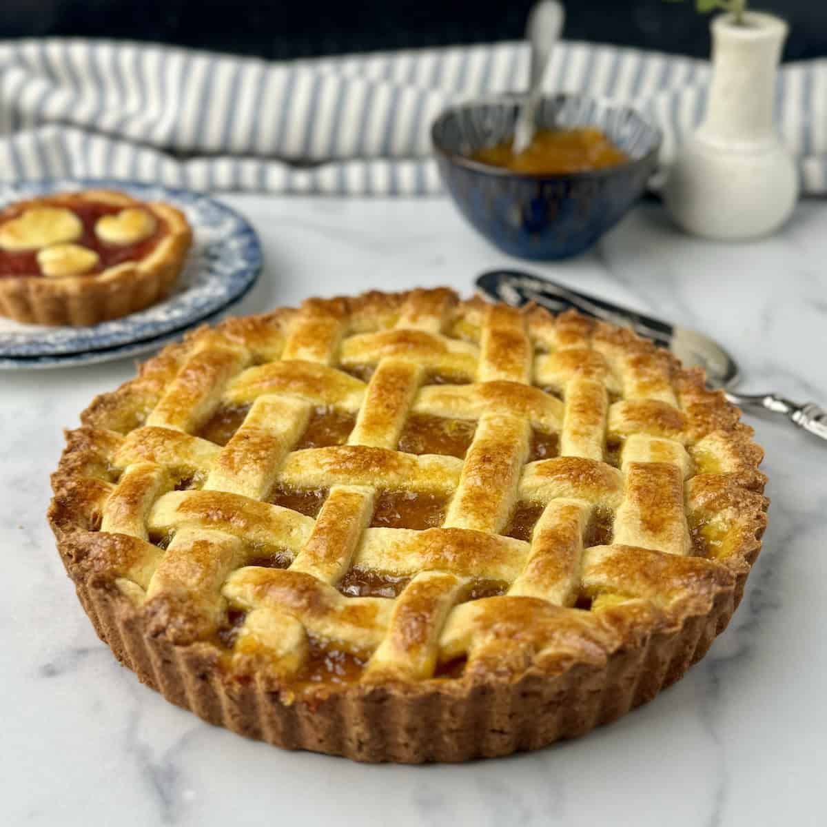 Pasta flora jam tart with one slice out next to mini jam tarts and a bowl with strawberry jam.