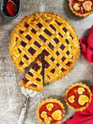 Pasta flora jam tart with one slice out next to mini jam tarts and a bowl with strawberry jam.