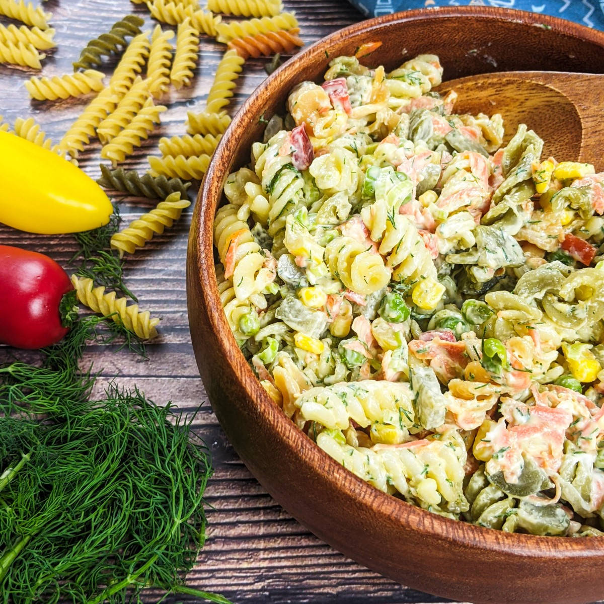 Fusilli pasta salad served in wooden bowl next to fresh dill, peppers and tricolore fusilli pasta.